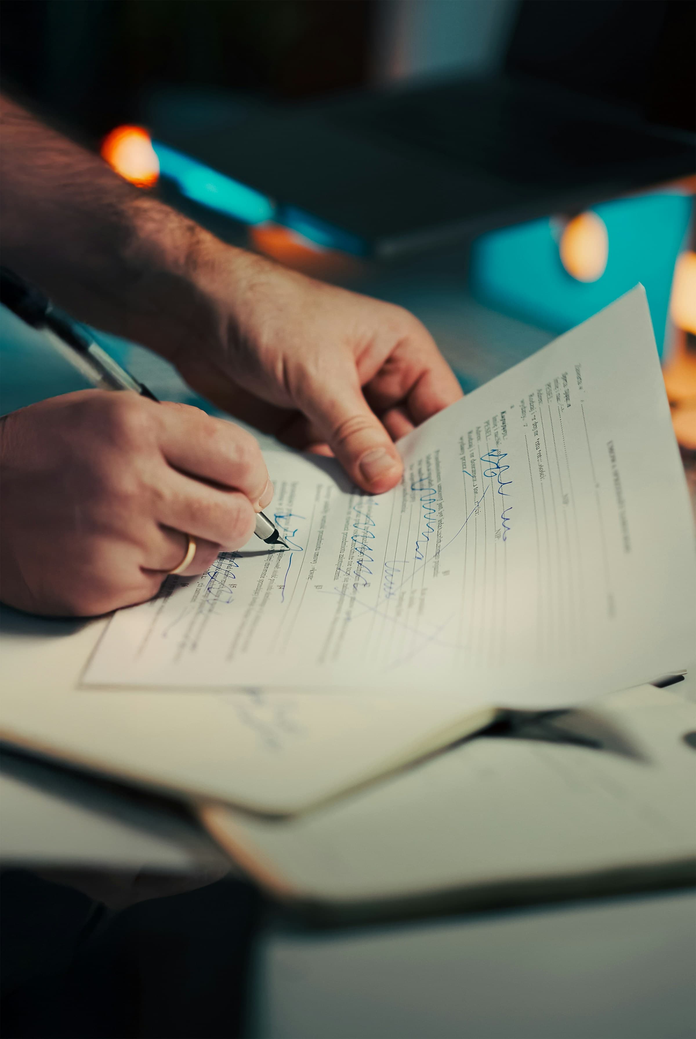 Hands signing and marking up a legal document on a dim desk with warm lights in the background.